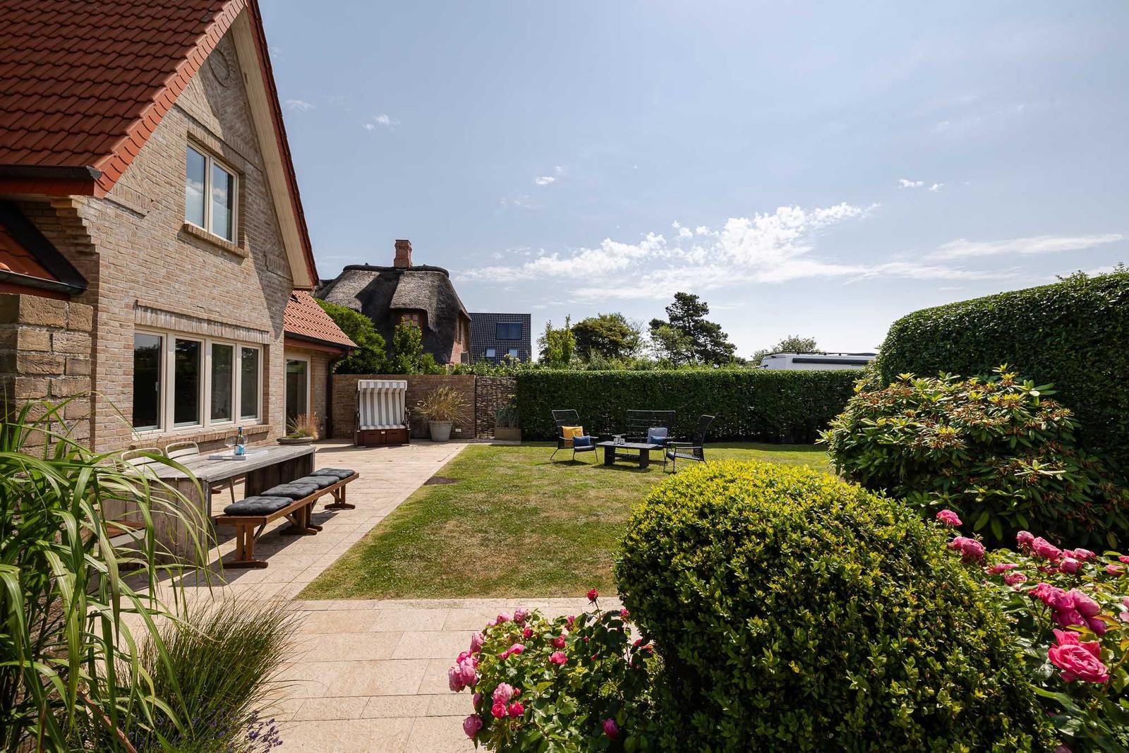 Hinterhof mit Terrasse, Gartenmöbeln und Blumenbeeten. Haus mit Ziegeldach und Hecke im Hintergrund.