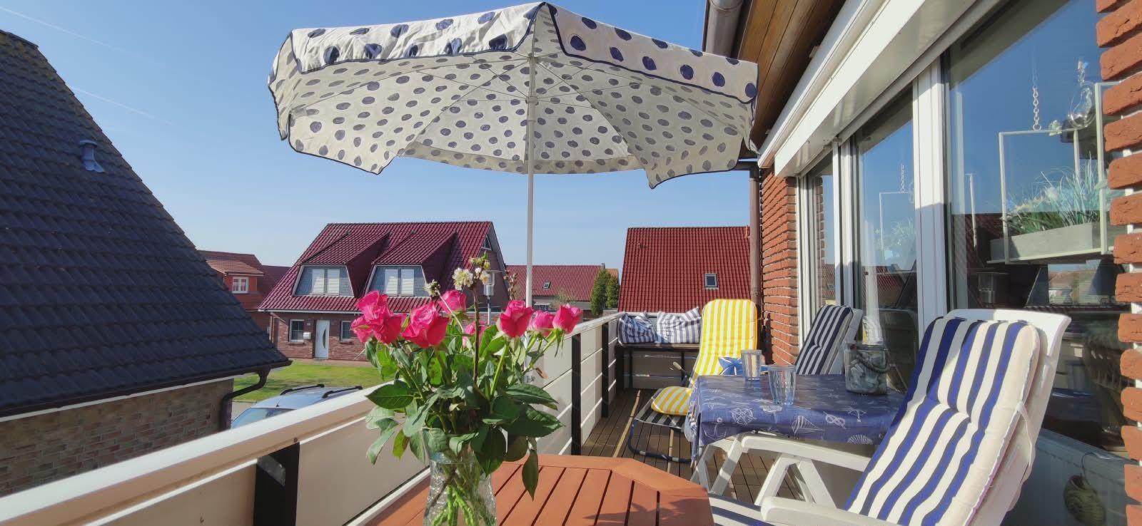 Terrasse mit Sonnenschirm, Stühlen und Blumen. Blick auf rote Dächer und grüne Wiesen.