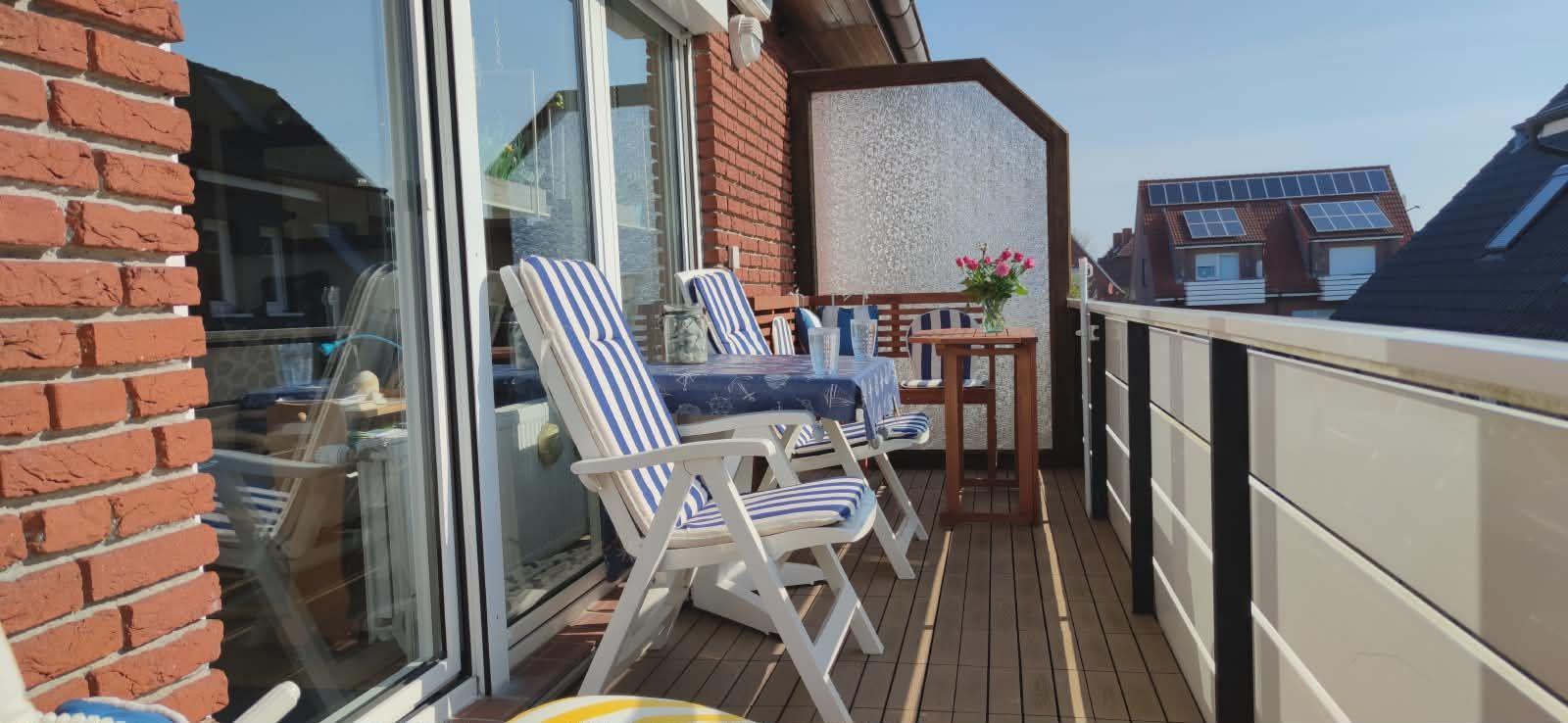 Terrasse mit blau-weißen Stühlen, Tisch und Blumen. Holzdielen und rote Ziegelmauer.