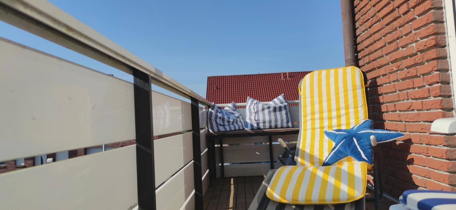 Terrasse mit gelb-weißem Liegestuhl und blau-weißem Kissen, roter Ziegelwand und Blick auf Dach