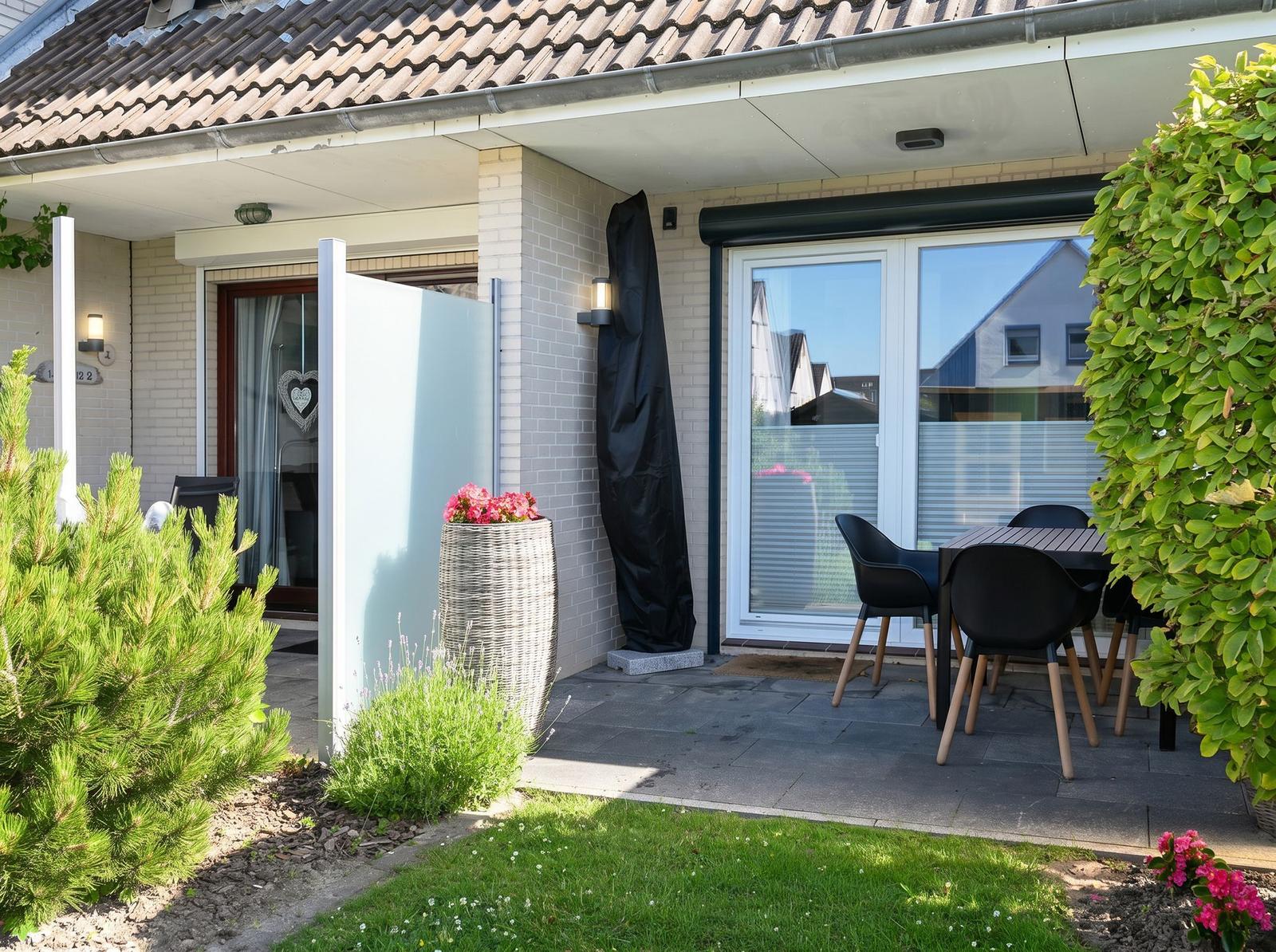 Terrasse mit Tisch und Stühlen, Pflanzen und Gartentür. Eingangstür und Fenster sichtbar.