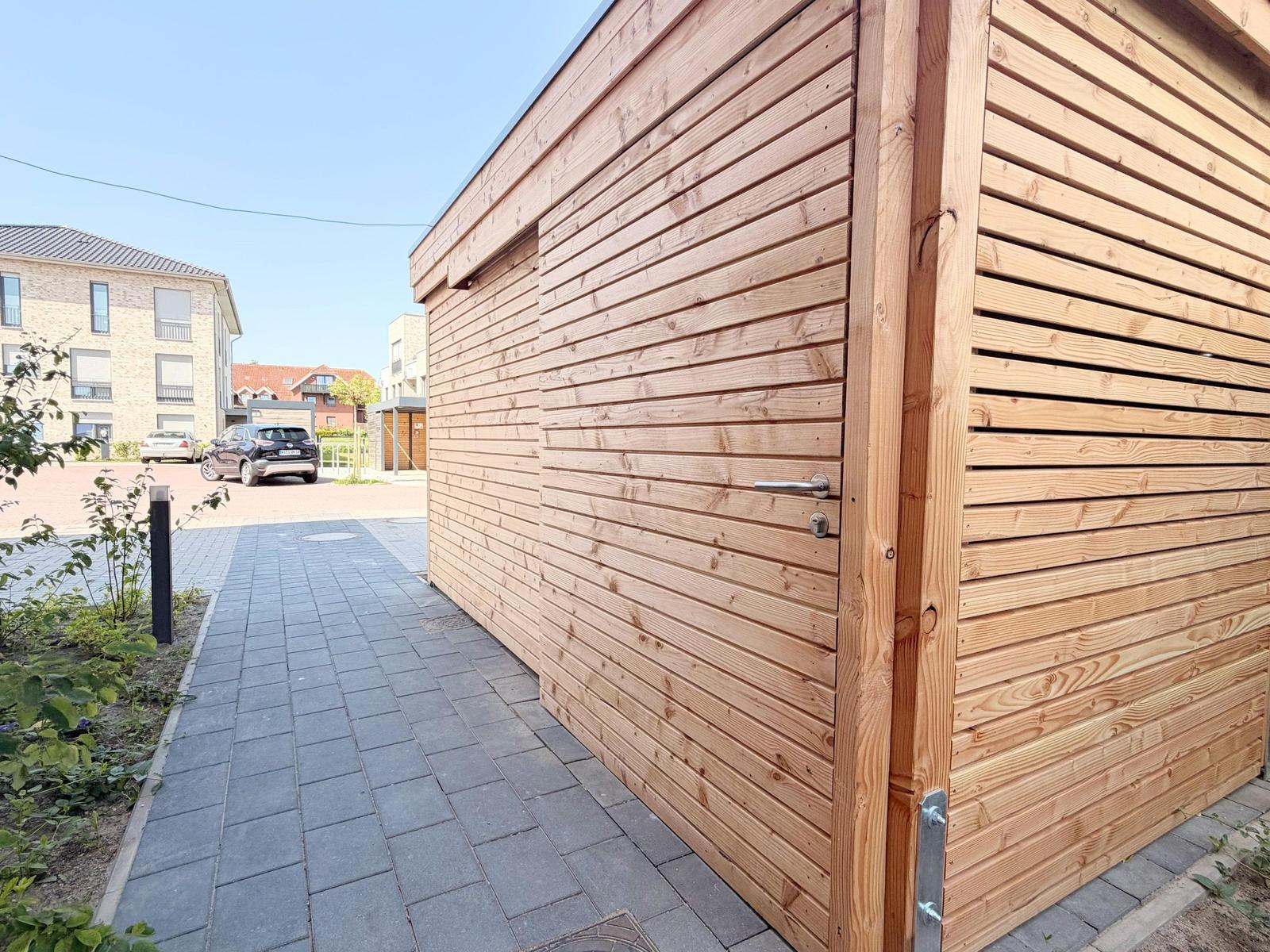 Holzgarage mit Tür und gepflastertem Weg. Im Hintergrund sind Gebäude und ein Auto sichtbar.