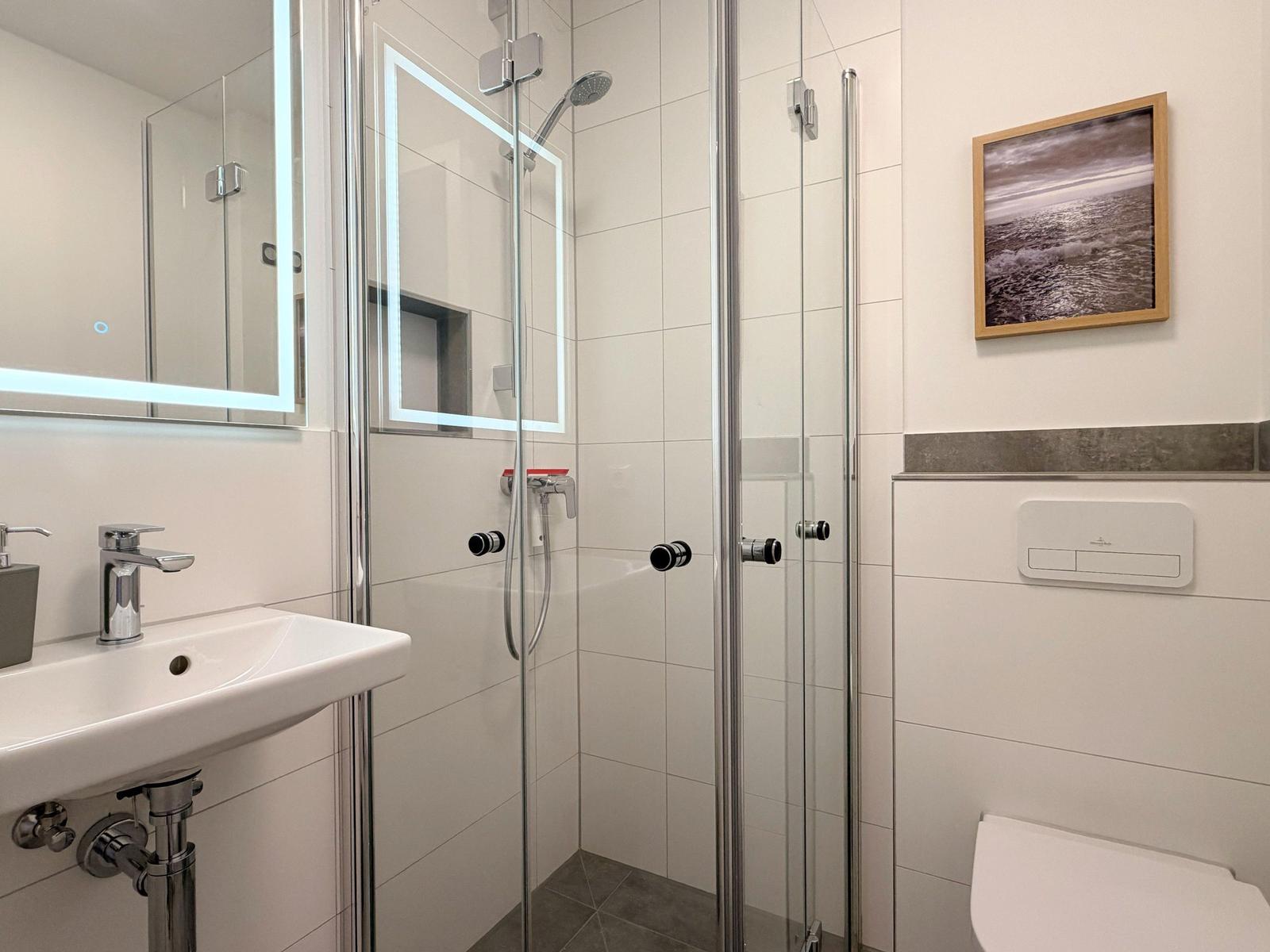 Badezimmer mit Dusche, Waschbecken und Toilette. Spiegel mit Beleuchtung und Wandbild.