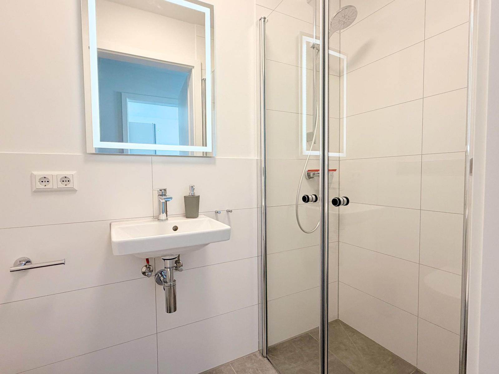 Badezimmer mit Dusche, Waschbecken und beleuchtetem Spiegel.