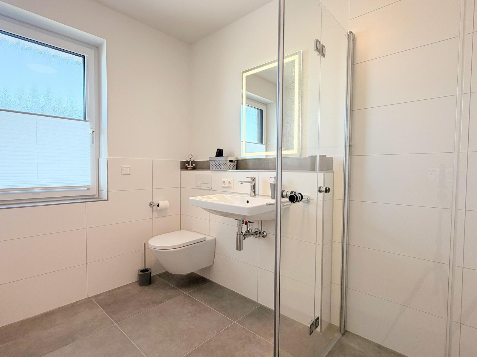 Badezimmer mit Dusche, Waschbecken und Toilette. Fenster mit Jalousie.