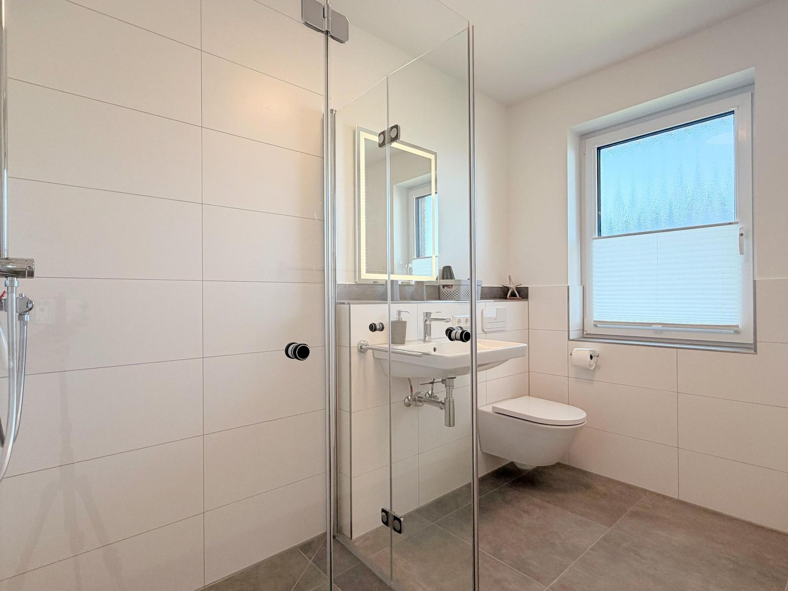 Badezimmer mit Dusche, Waschbecken und Toilette. Fenster mit Jalousie.