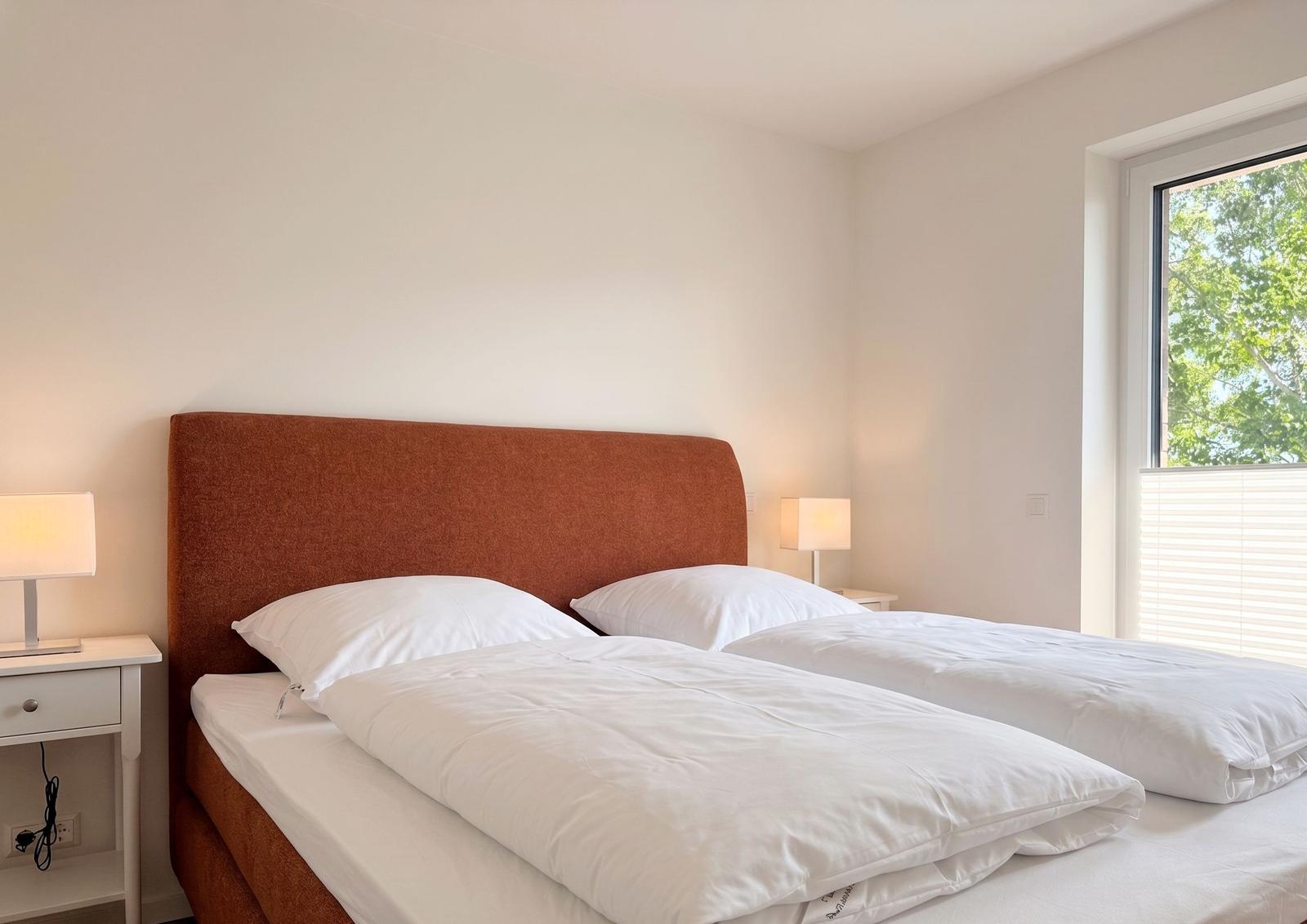 Schlafzimmer mit Doppelbett, orangefarbenem Kopfteil und Fenster mit Blick auf Bäume.