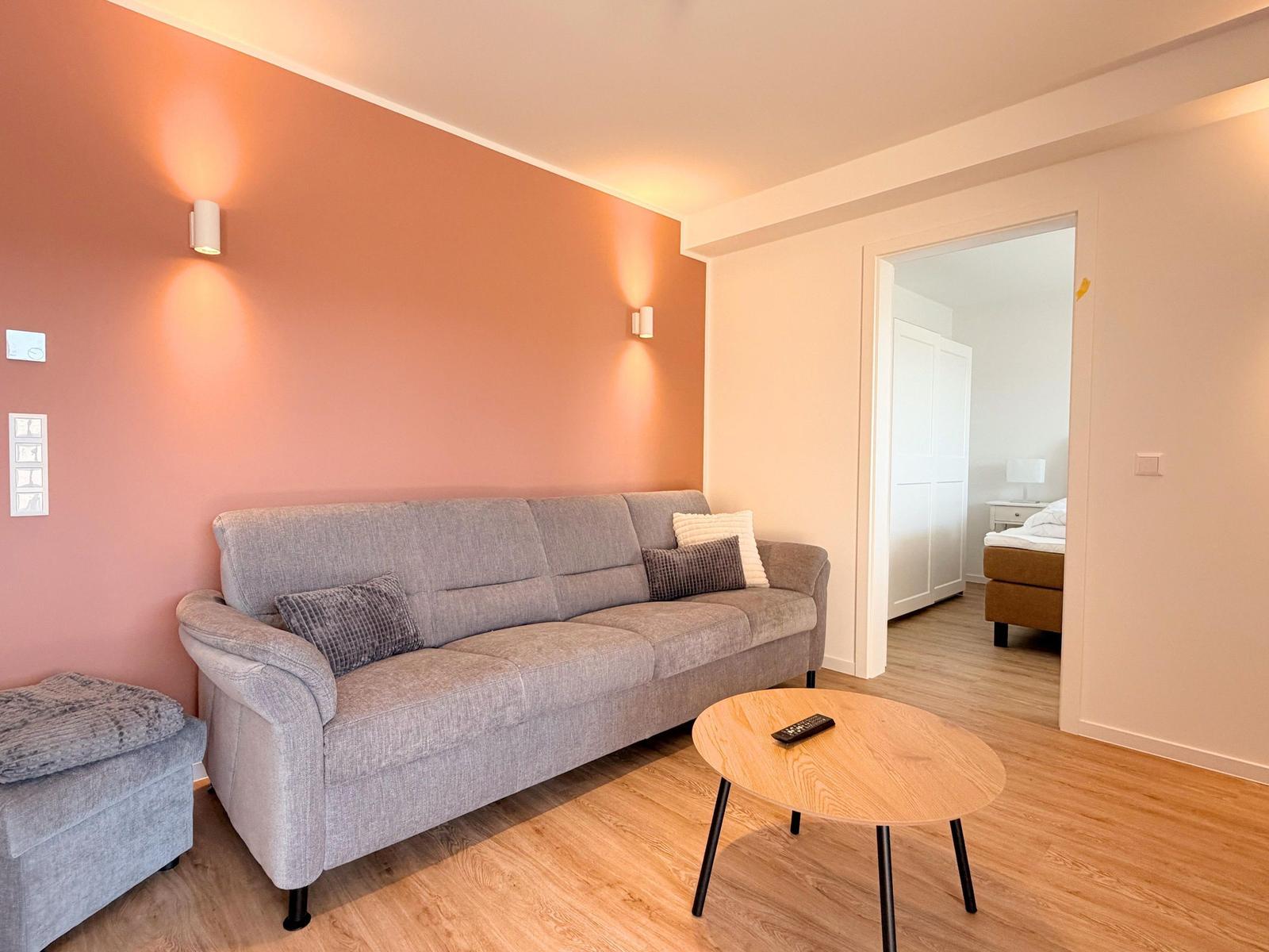 Wohnzimmer mit grauem Sofa, runden Tisch und Tür zum Schlafzimmer.