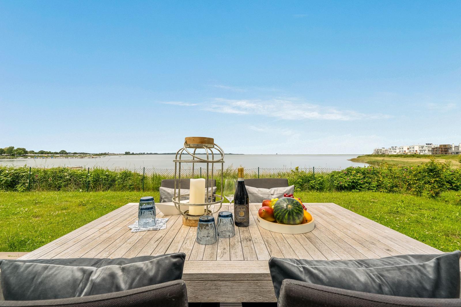 Terrasse mit Tisch, Stühlen und Blick auf Wasser und Grün.