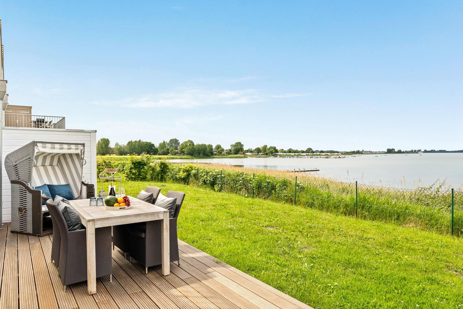 Terrasse mit Tisch, Stühlen und Strandkorb. Blick auf ruhiges Wasser und Grasfläche.