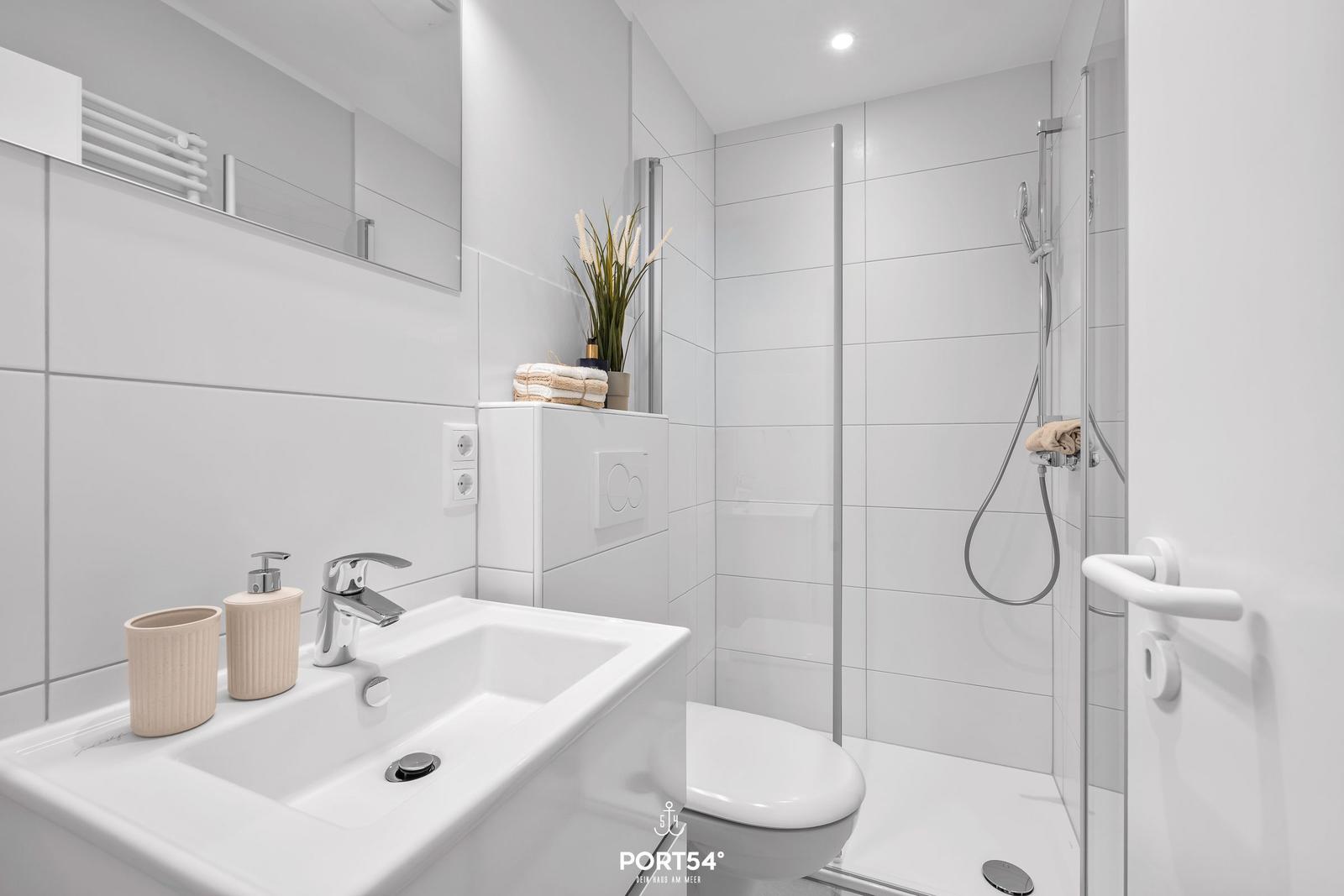 Badezimmer mit Dusche, Waschbecken und Toilette. Weiße Fliesen und moderne Einrichtung.