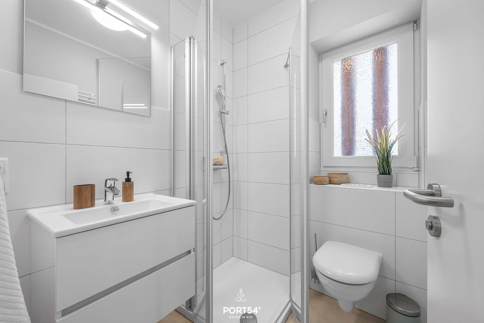 Badezimmer mit Dusche, Waschbecken und Toilette. Fenster mit Pflanze.