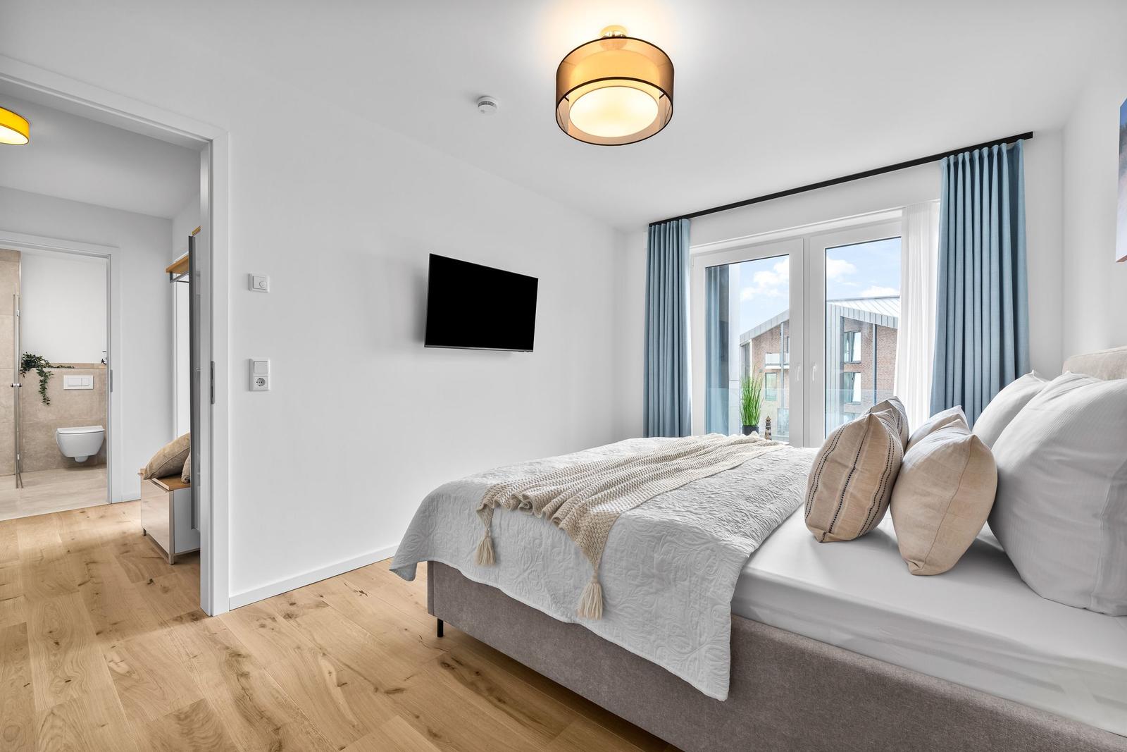 Schlafzimmer mit Bett, TV und Blick auf Balkon. Holzfußboden und blaue Vorhänge.