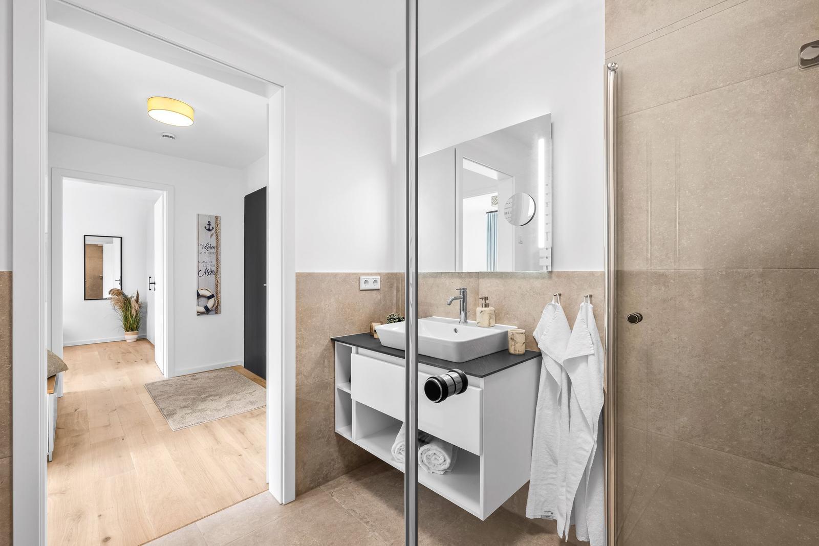 Badezimmer mit Dusche, Waschtisch und Spiegel. Blick in den Flur mit Holzboden.