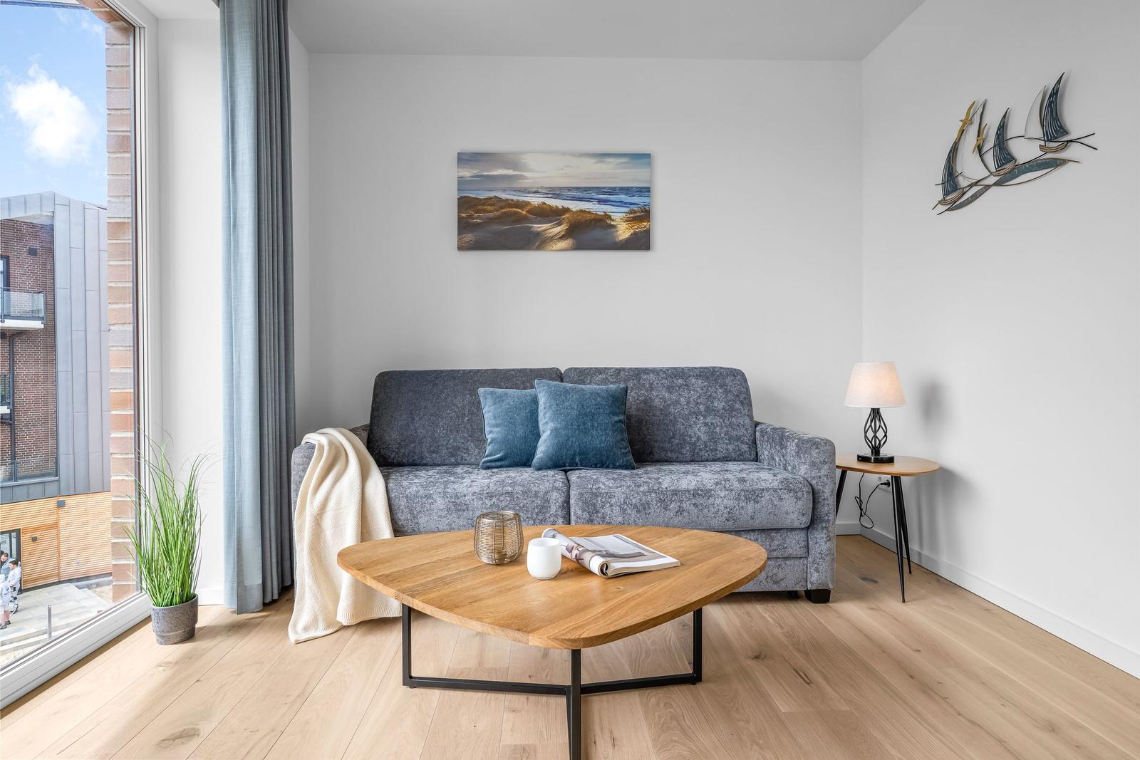Ein Wohnzimmer mit grauem Sofa, Holztisch und Wandbild. Große Fenster mit Blick auf Gebäude.
