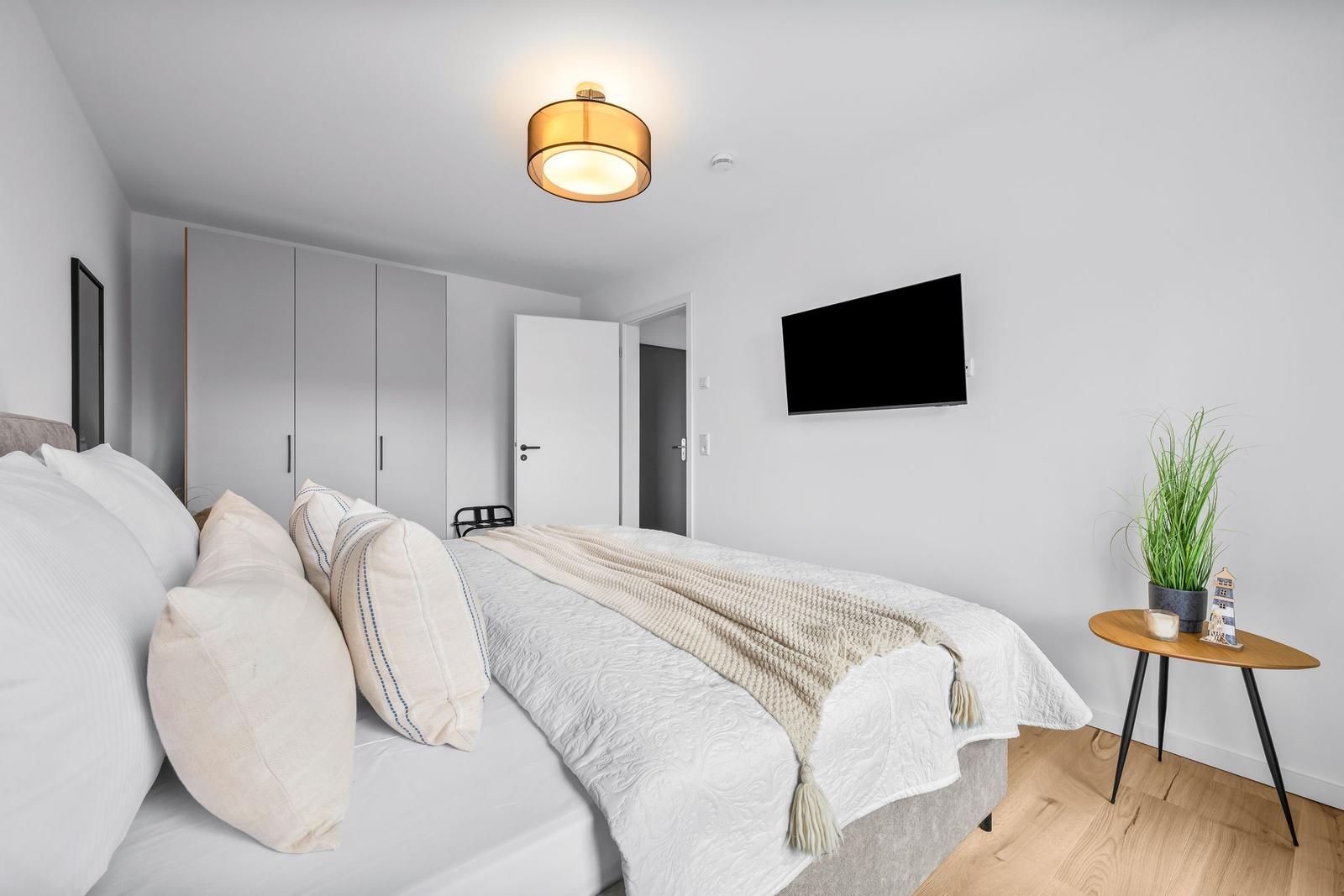 Schlafzimmer mit Bett, Kleiderschrank, Fernseher und Pflanze auf Tisch.
