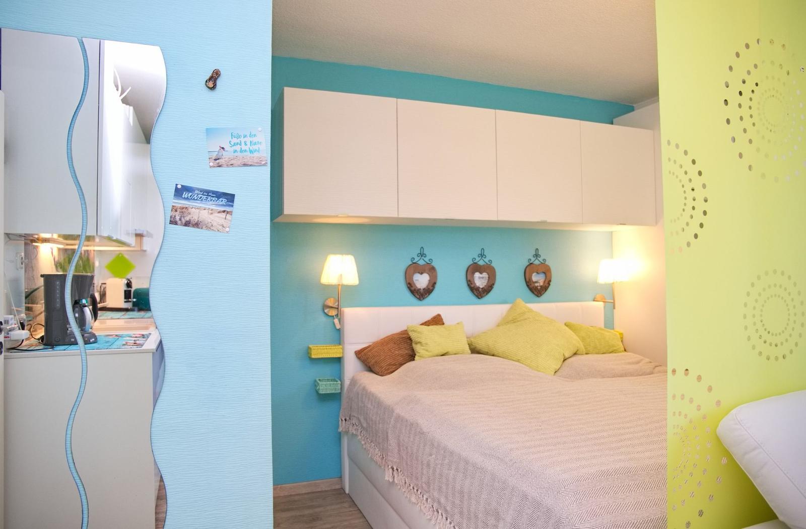 Schlafzimmer mit Bett, Nachttisch und Wandregalen. Küchenbereich mit Spüle und Kühlschrank.