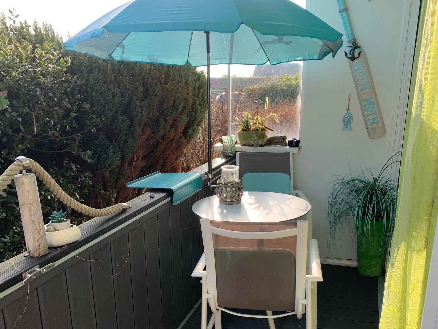 Terrasse mit Tisch, Stühlen und blauem Sonnenschirm. Pflanzen und Dekoration an der Wand.