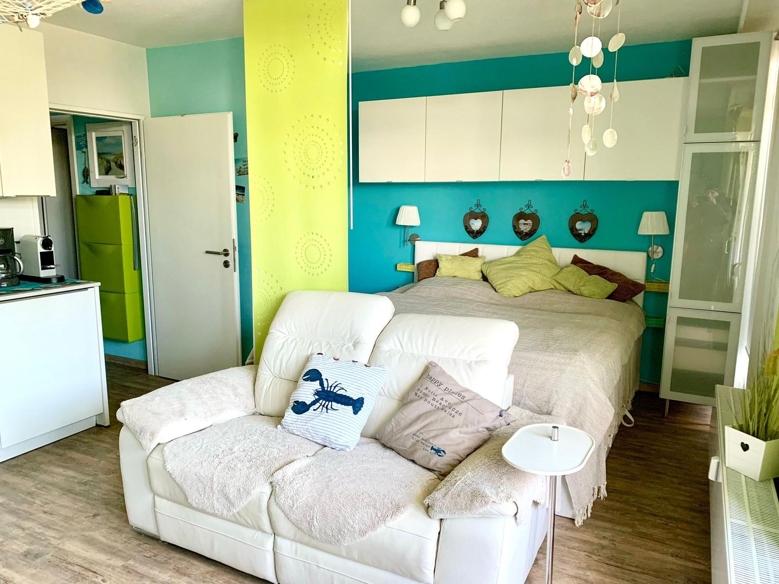 Schlafzimmer mit weißem Sofa, Bett und farbigen Wänden. Küche im Hintergrund.