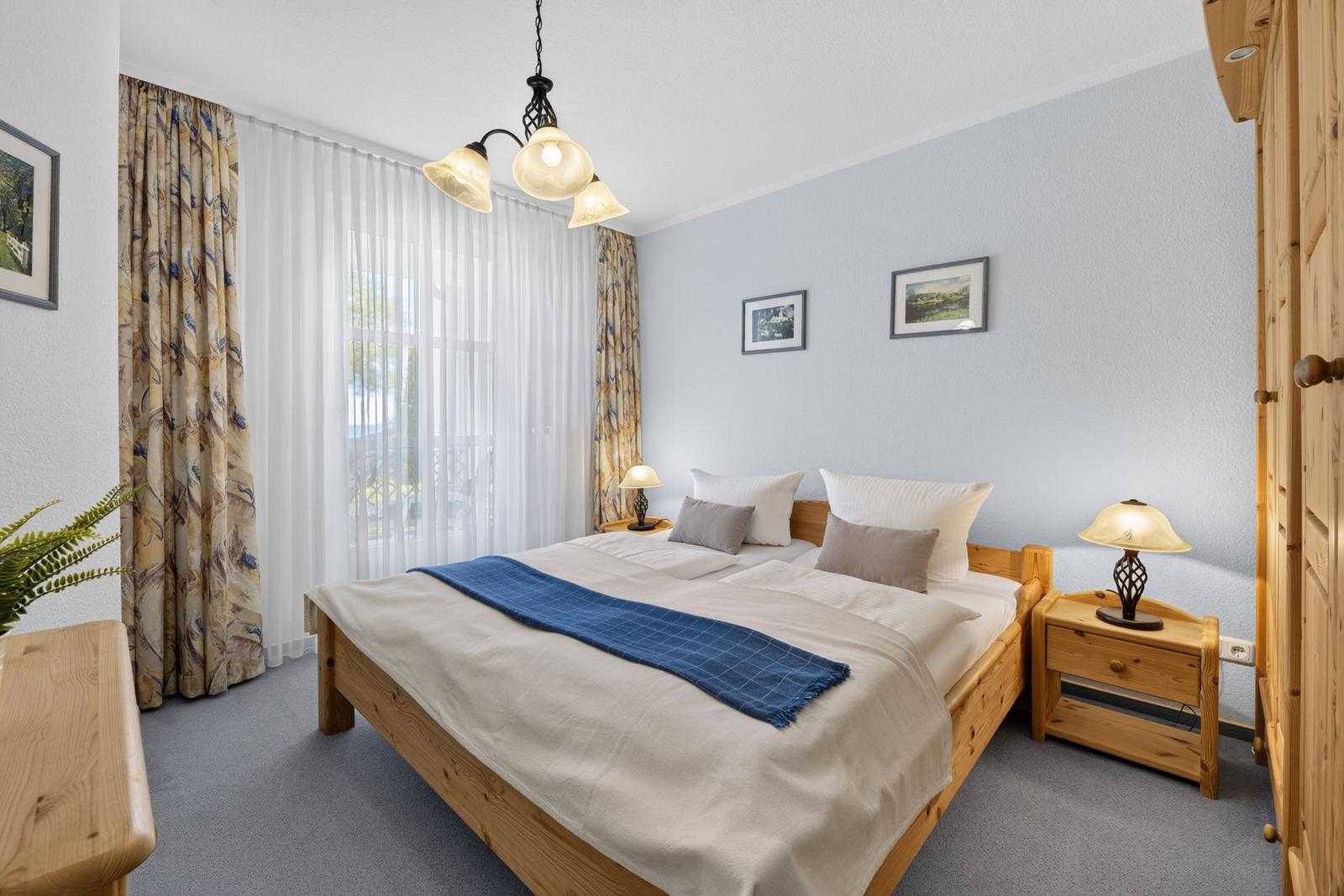 Schlafzimmer mit Holzbett, Nachttisch und Gardinen vor Fenster.