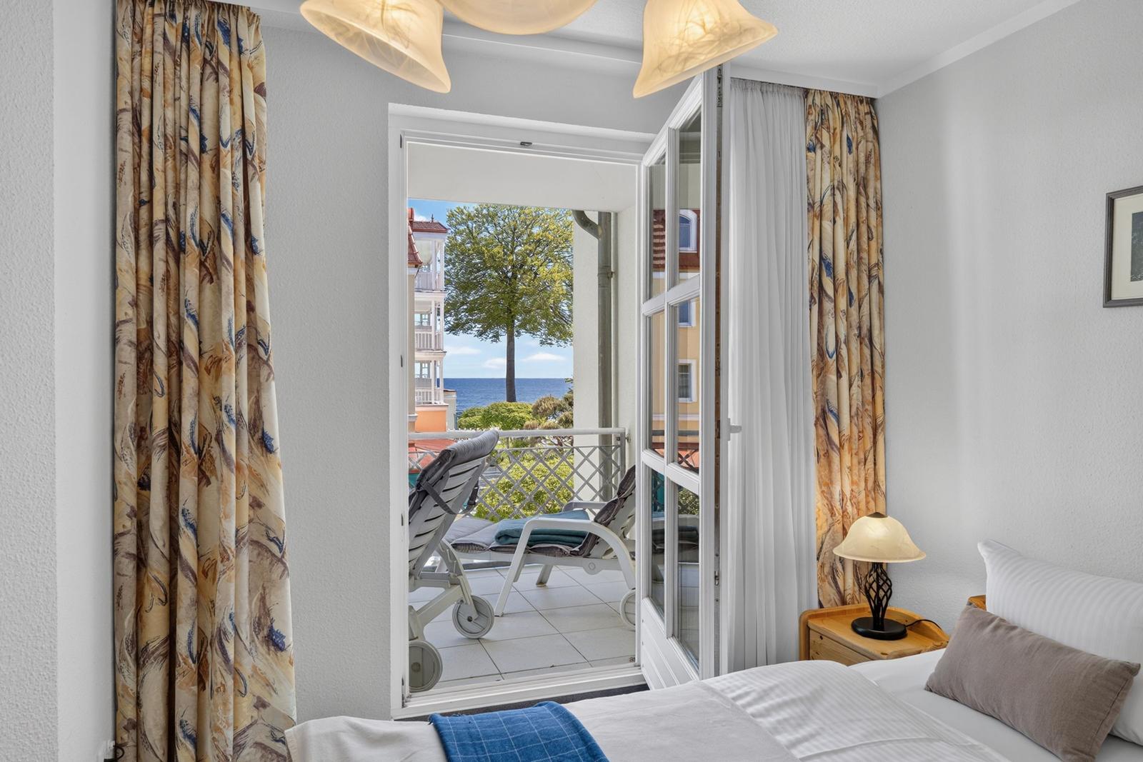 Zimmer mit Blick auf Balkon und Meer. Vorhänge, Bett und Lampe sichtbar.