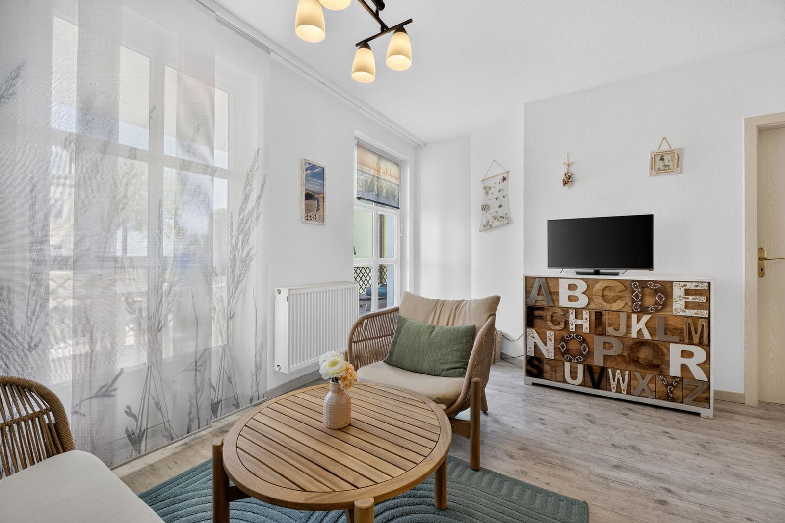 Wohnzimmer mit Sitzgelegenheit, TV und Holztisch. Fenster mit weißen Vorhängen.