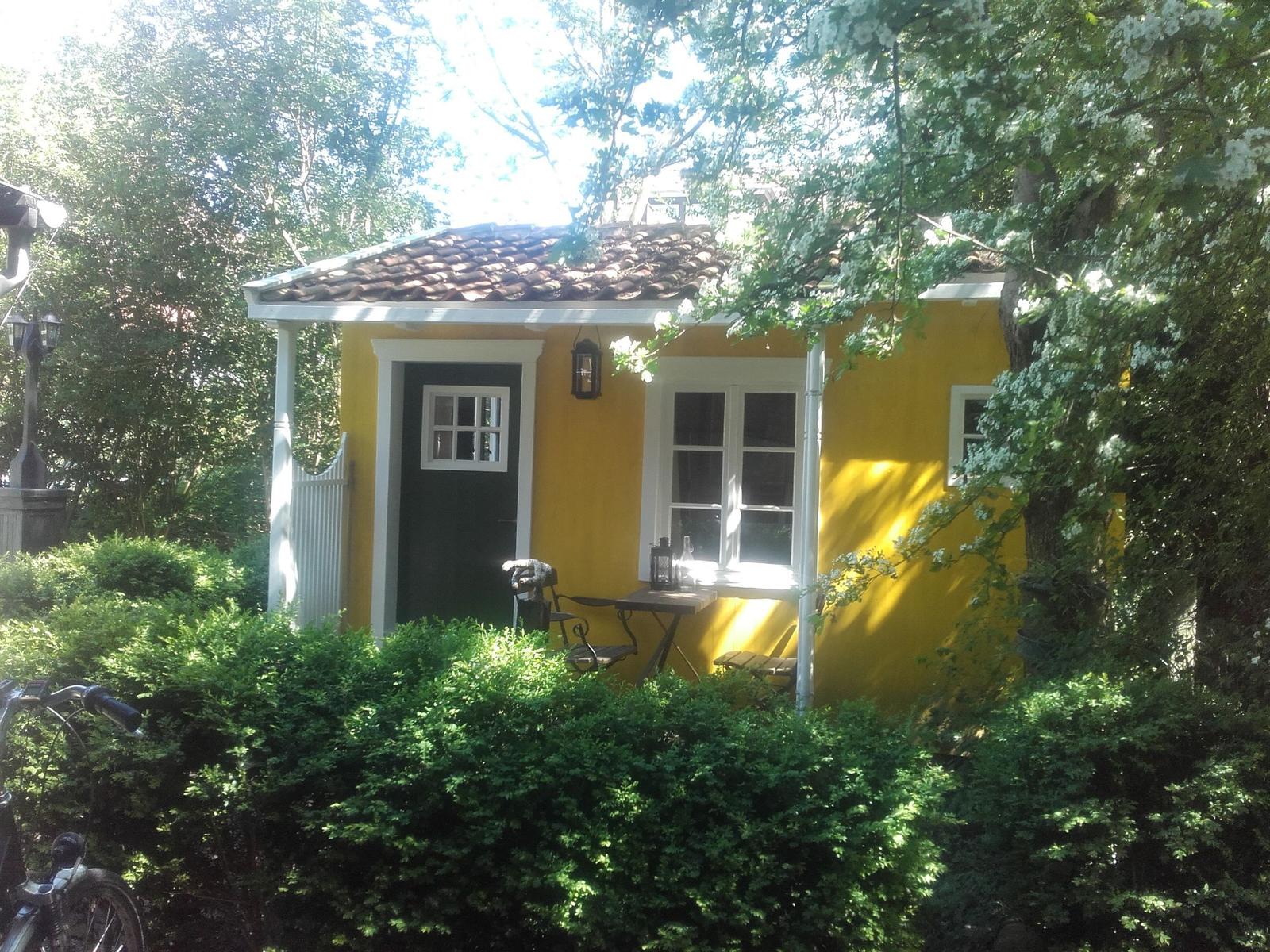 Gelbes Häuschen mit grüner Tür, Terrasse und umgebenden Bäumen und Sträuchern.