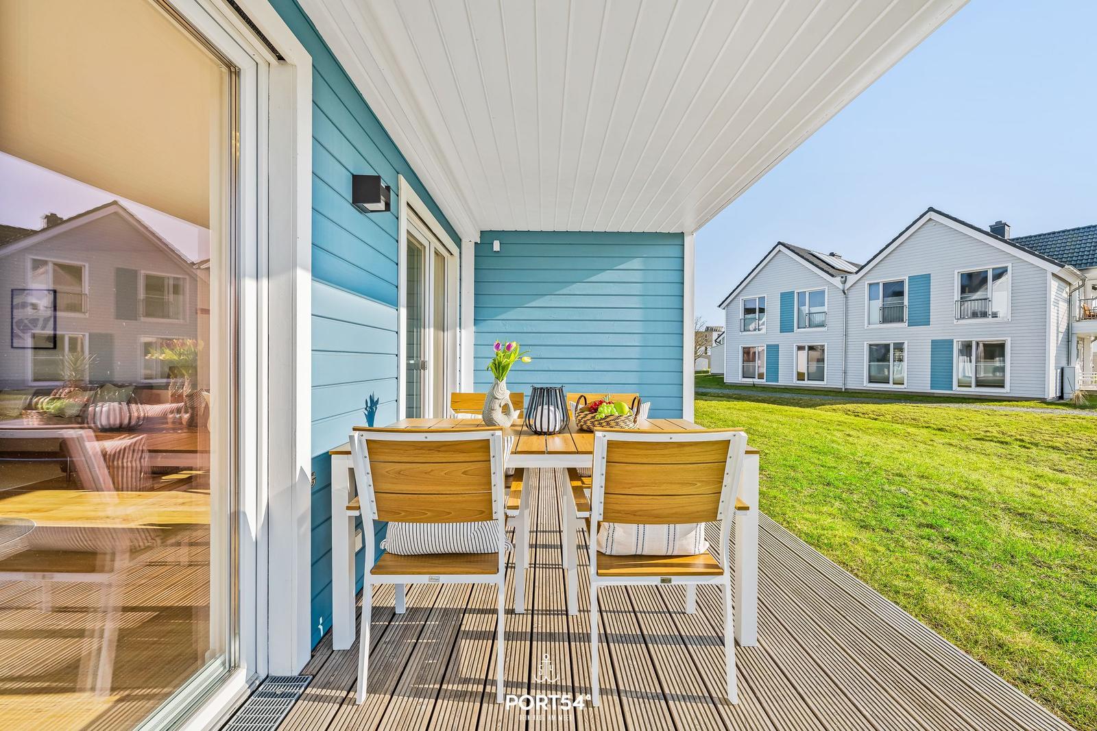 Terrasse mit Tisch und Stühlen, blauer Holzaufbau, Grünfläche und Nachbarhäuser im Hintergrund.