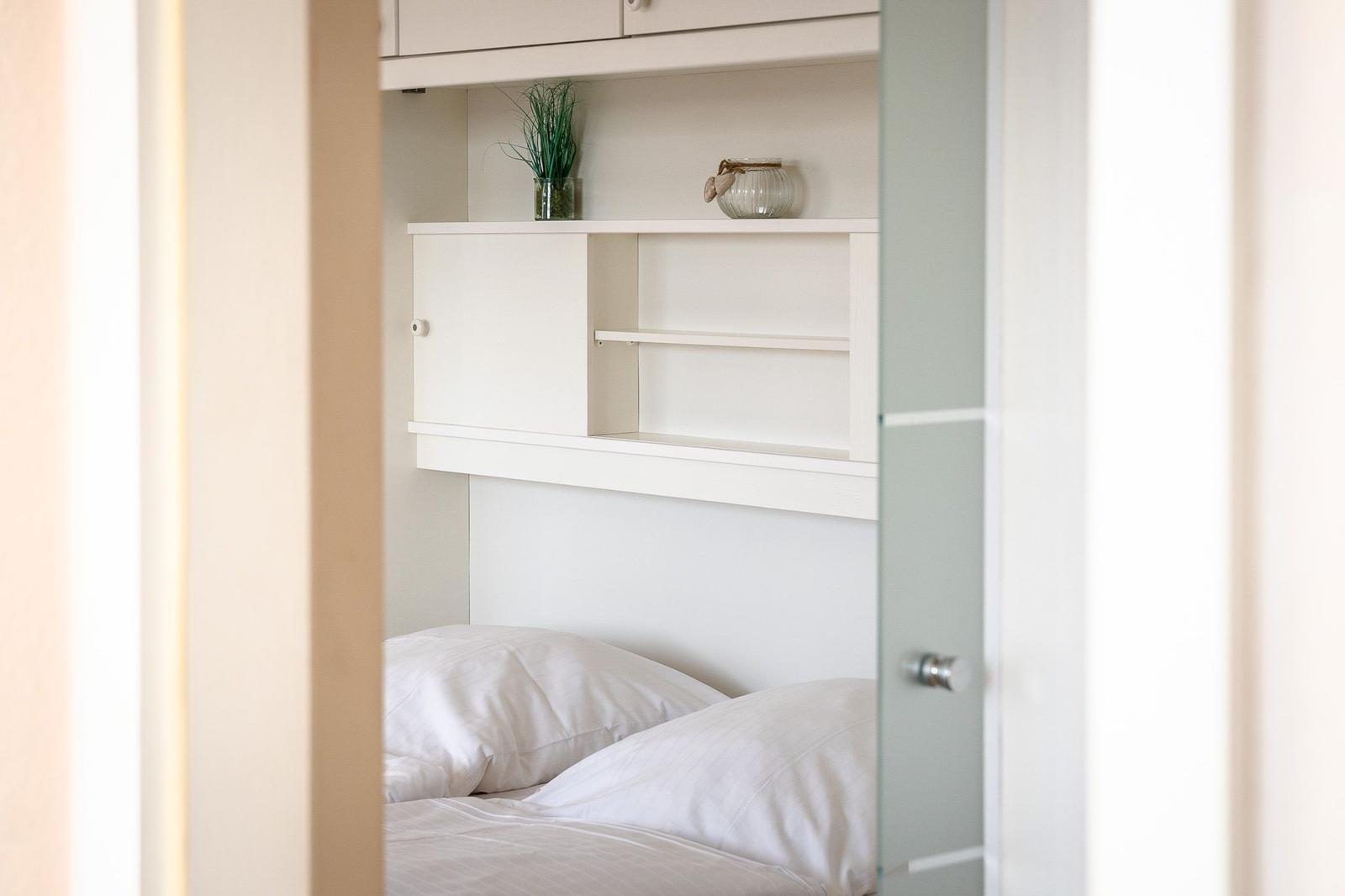 Bedroom with white bed and integrated cabinet with shelf and plant.