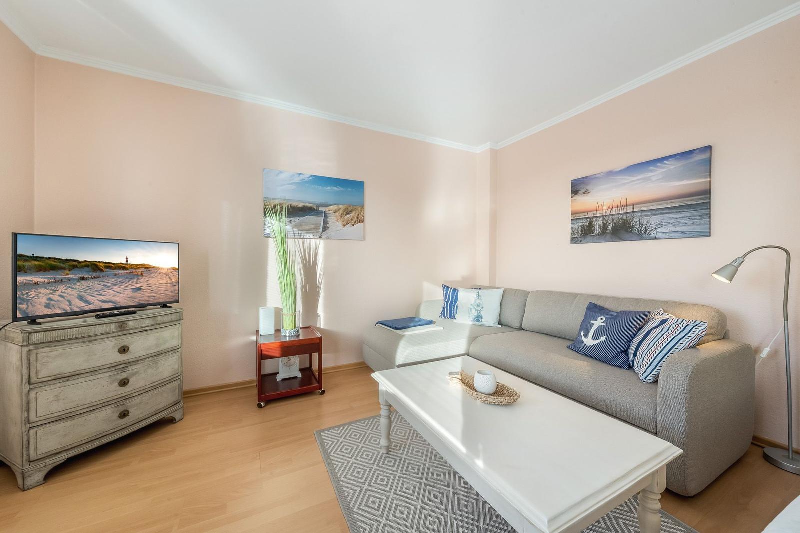 Living room with sofa, coffee table, and TV. Wall art shows beach landscapes.