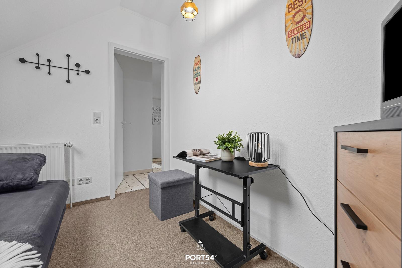 Schlafzimmer mit Bett, Schreibtisch und Kleiderschrank. Wanddekoration und Pflanze sichtbar.