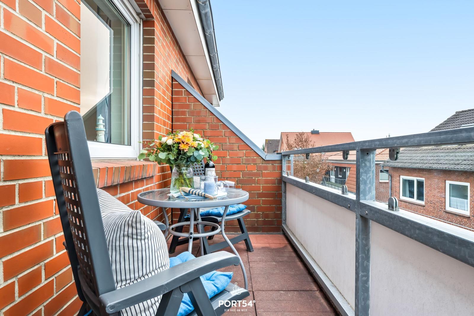 Balkon mit Tisch, Stühlen und Blumenarrangement. Blick auf Wohnhäuser und Himmel.