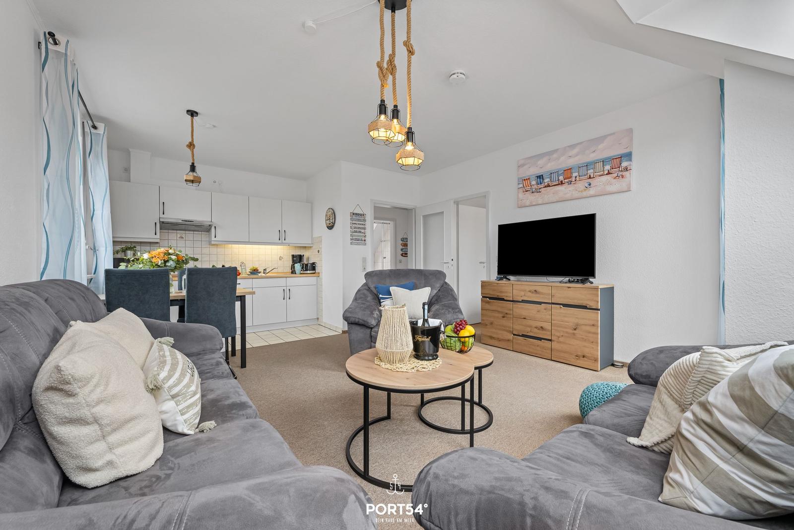 Wohnzimmer mit Sofa, Couchtisch und TV. Küche im Hintergrund.