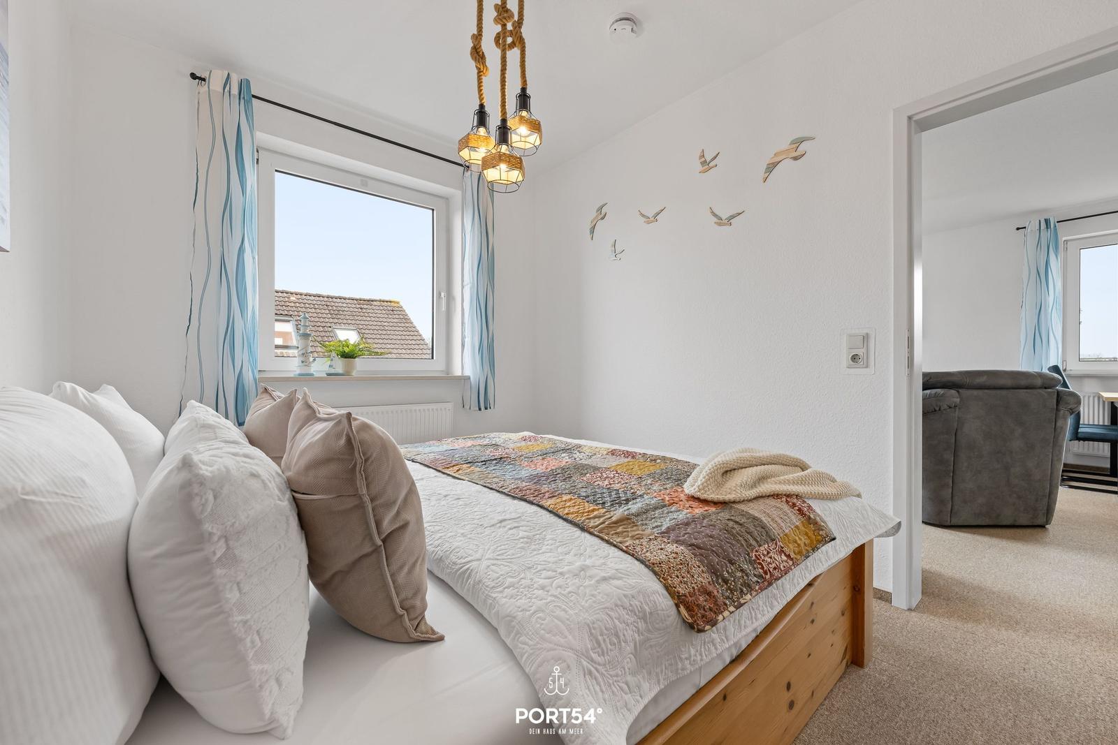 Schlafzimmer mit Bett, Fenster und Wanddekoration. Durchgang zum Wohnbereich.