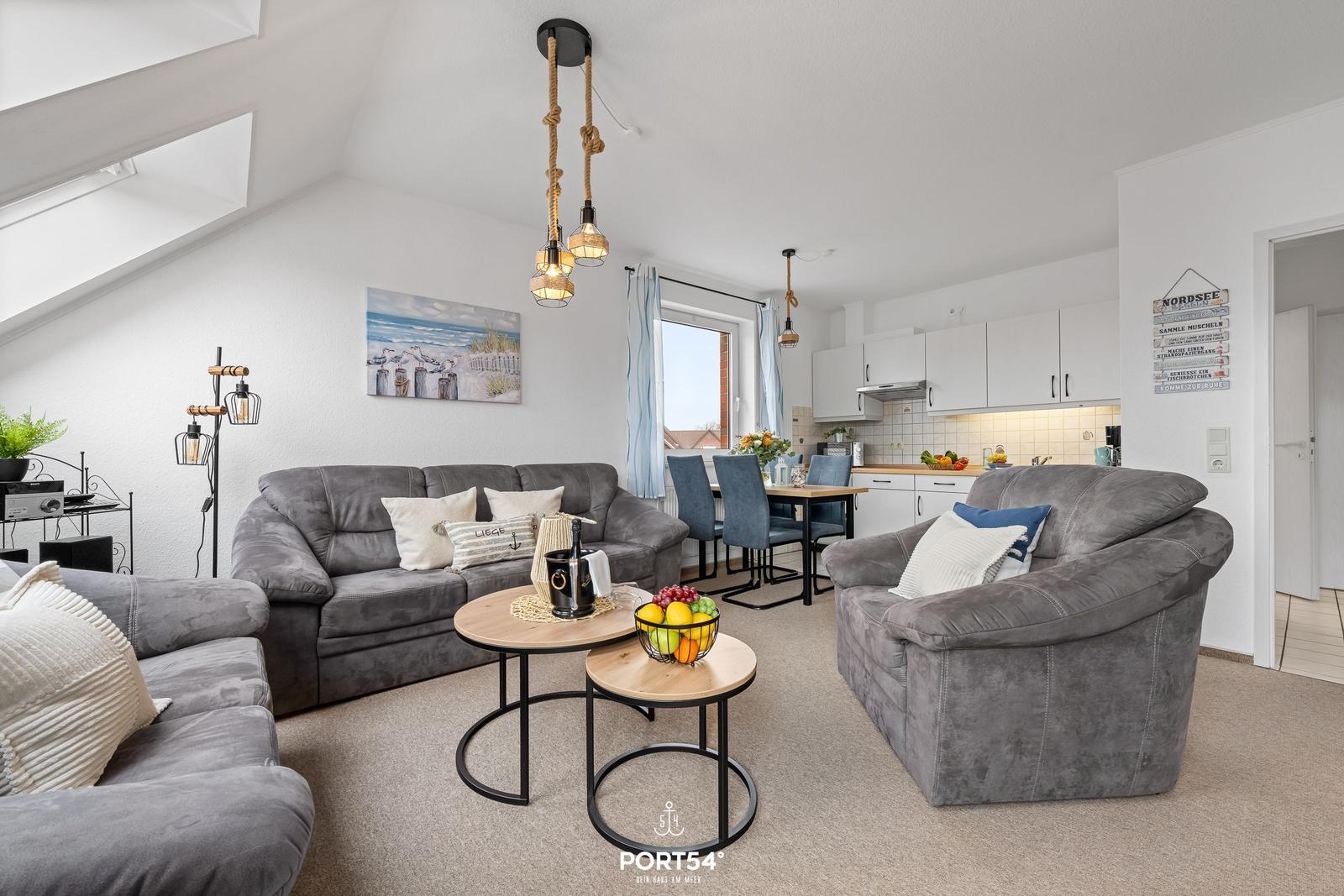 Wohnzimmer mit Sofa, Sessel und Essbereich. Hängelampe, Fenster und Küchenzeile sichtbar.