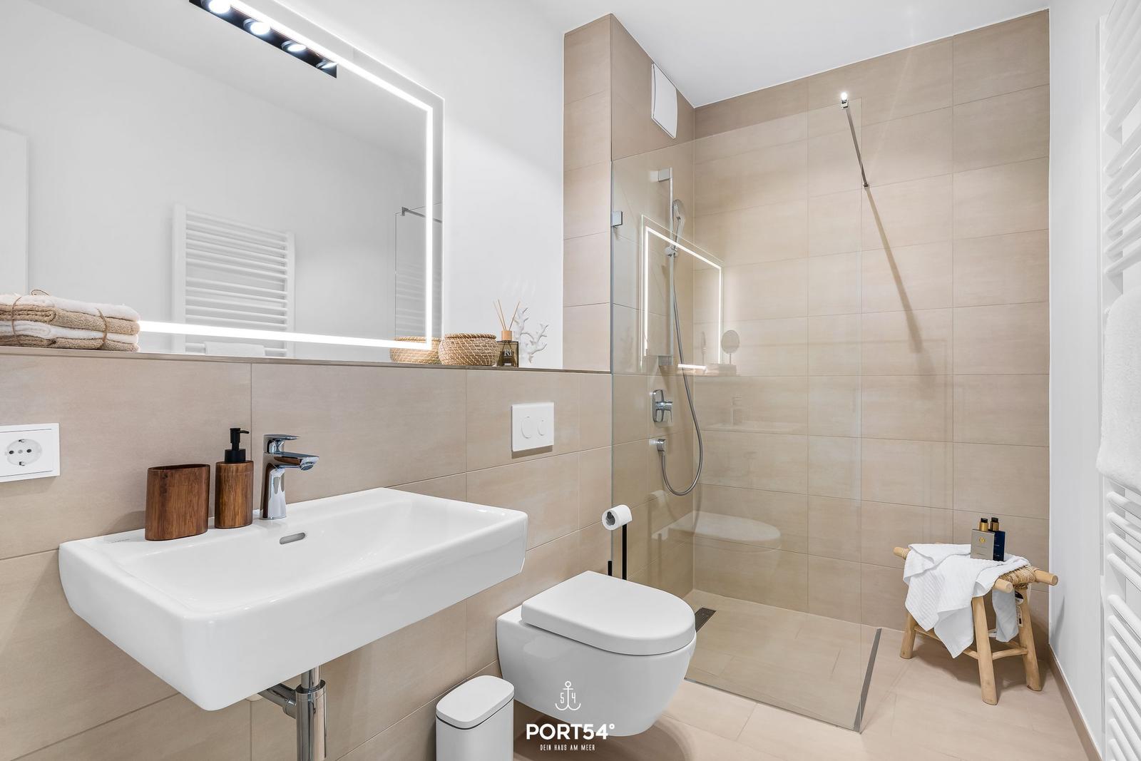 Bathroom with shower, sink and toilet. Lit mirror and wooden accessories.