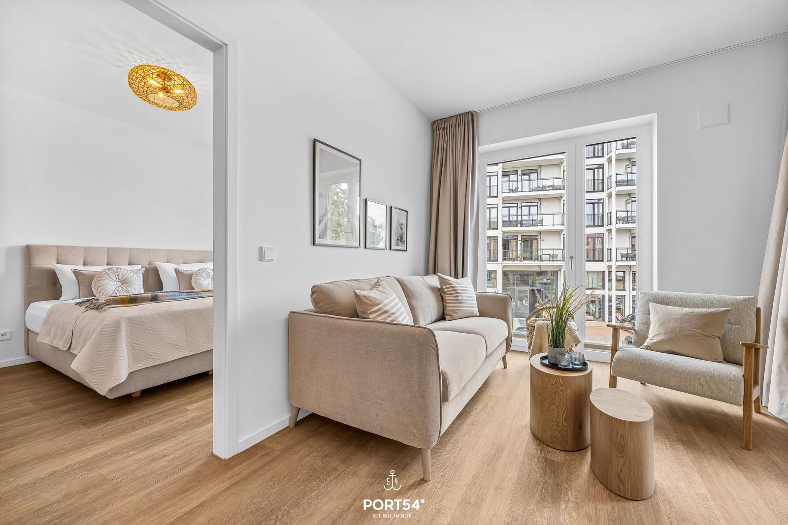 Living room with sofa, armchair, and balcony view. Adjacent room with bed and duvet.