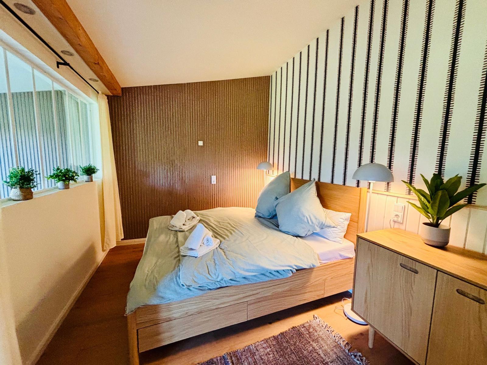 Schlafzimmer mit Bett, Nachttisch und Pflanzen an Fensterbank.