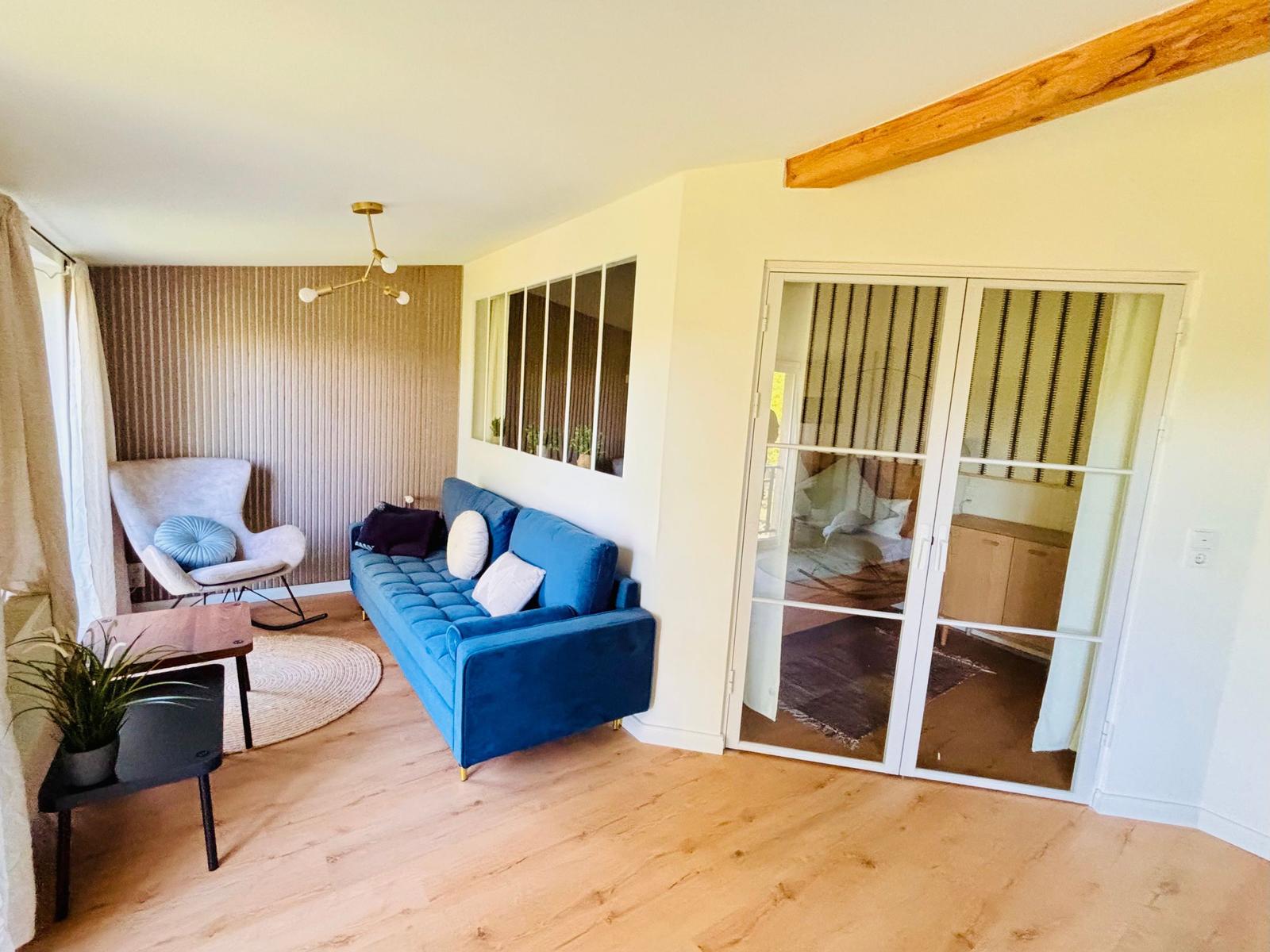Ein Wohnzimmer mit blauem Sofa, Sessel und Holzboden. Durchgang zu Schlafzimmer.