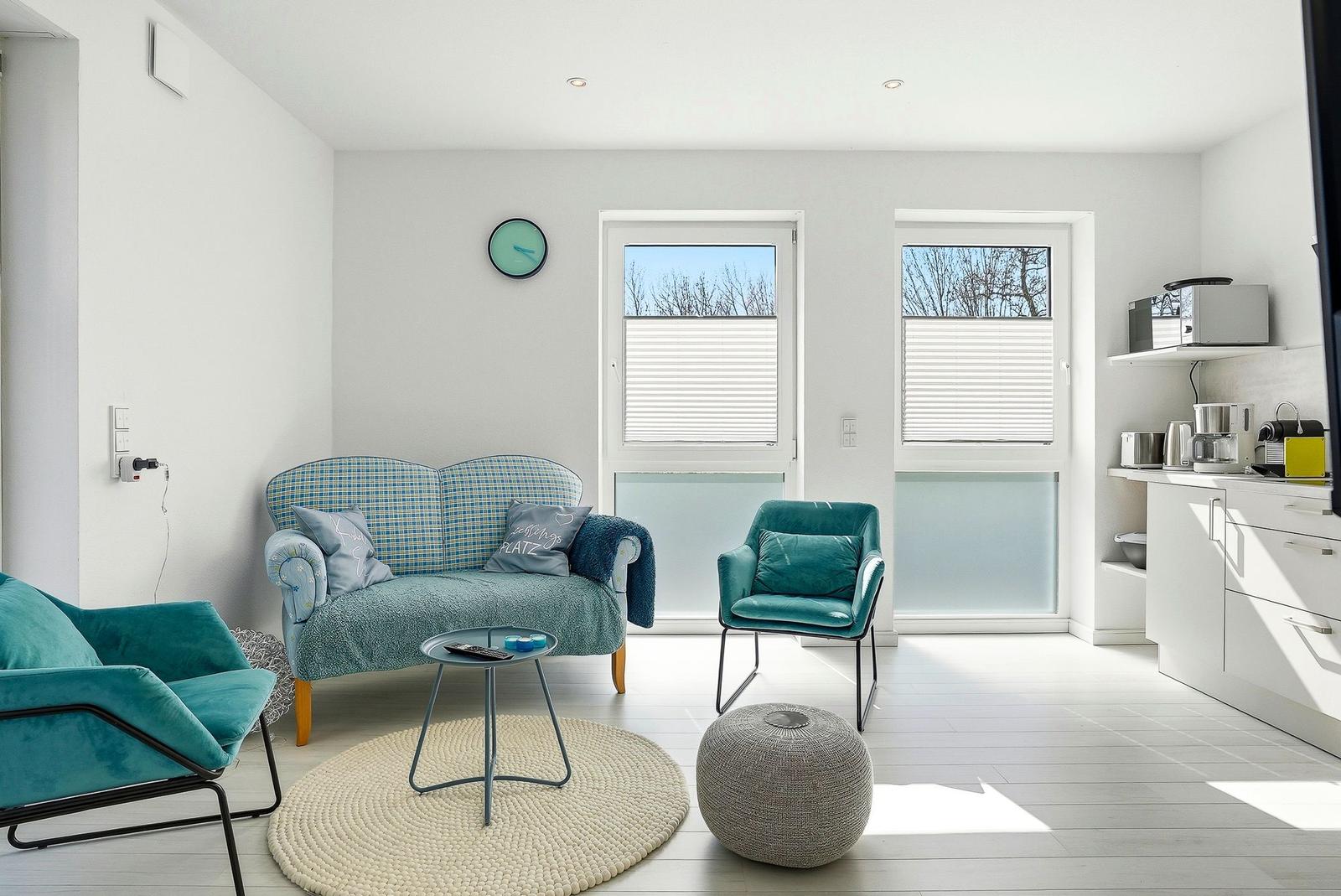 Wohnbereich mit Sofa, Sesseln und Küchenzeile. Fenster mit Jalousien und runder Teppich.