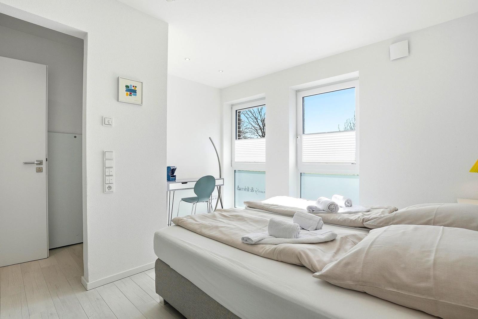 Schlafzimmer mit Bett, Schreibtisch und Fenster. Weiße Wände und Holzboden.