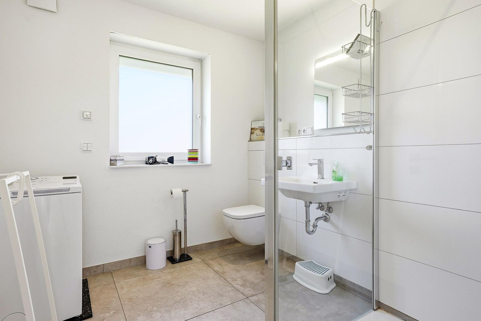 Badezimmer mit Dusche, Waschbecken und Toilette. Fenster, Waschmaschine und Spiegel sichtbar.