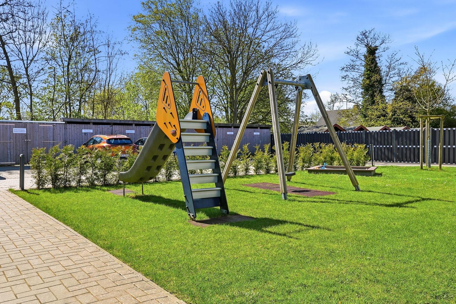 Grüner Garten mit Spielplatz, Rutsche und Schaukel. Hintergrund: Zaun und Bäume.