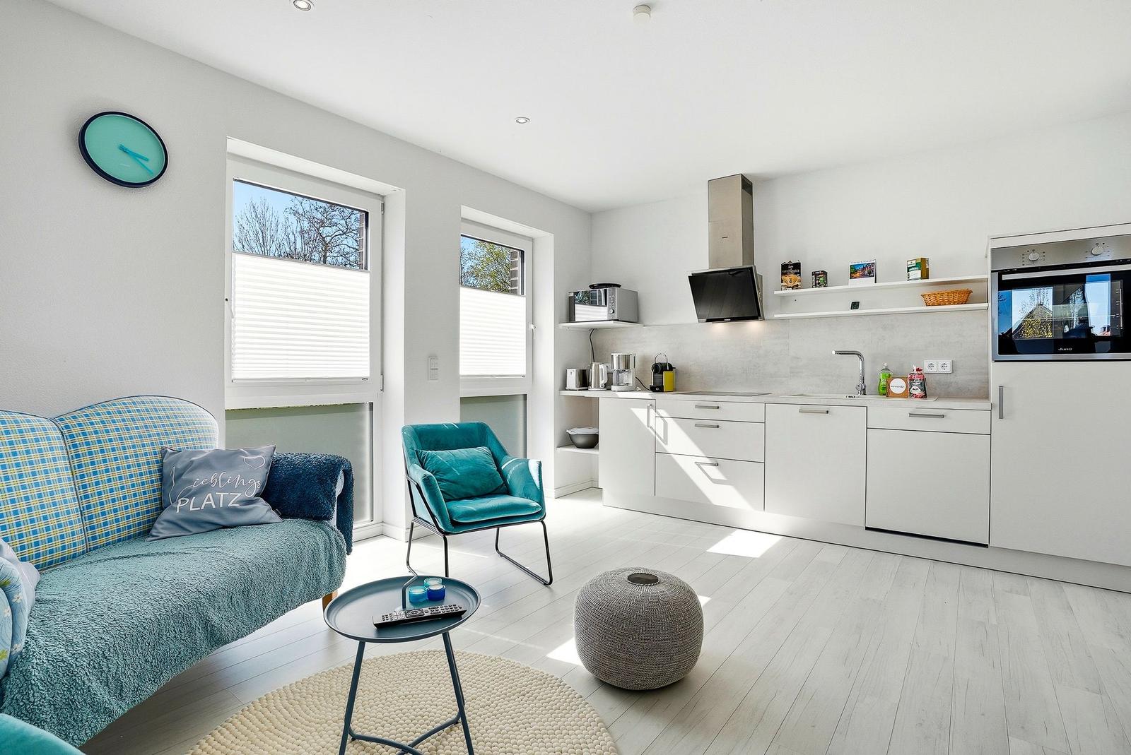 Wohnküche mit Sofa, Stuhl und Küchenschränken. Fenster mit Jalousien.
