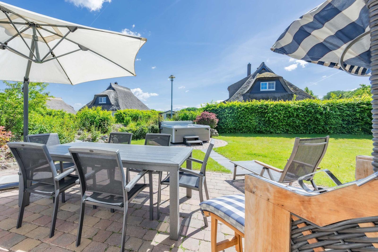 Terrasse mit Tisch, Stühlen und Sonnenschirmen. Im Hintergrund sind Häuser mit Strohdächern und ein Garten.