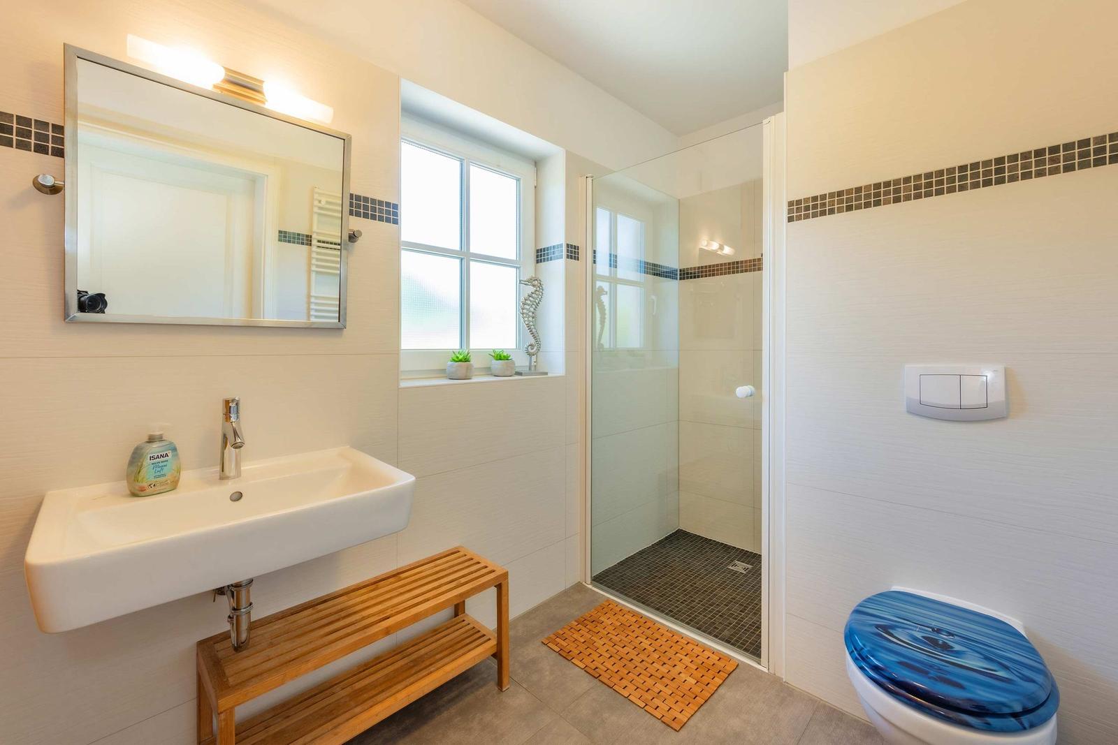 Badezimmer mit Dusche, Waschbecken und Toilette. Holzbank und Fenster mit Pflanzen.