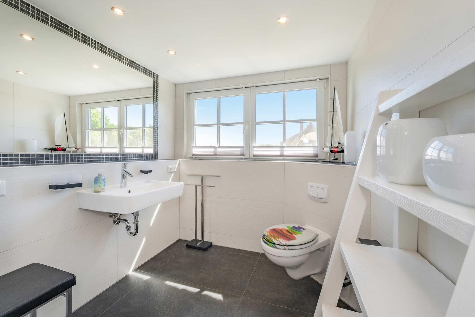 Badezimmer mit Waschbecken, Toilette und Fenster. Auf Regalen stehen weiße Behälter.
