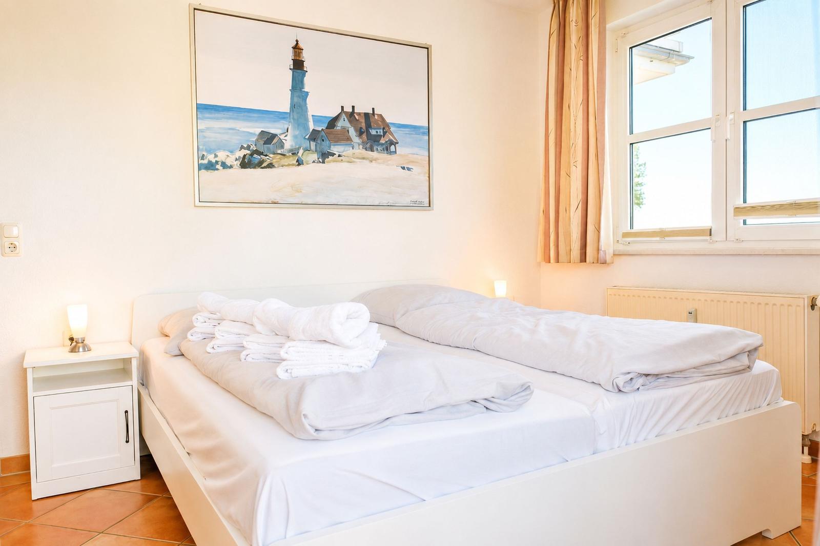 Schlafzimmer mit Bett, Nachttisch und Fenster. Wandbild zeigt Leuchtturm.