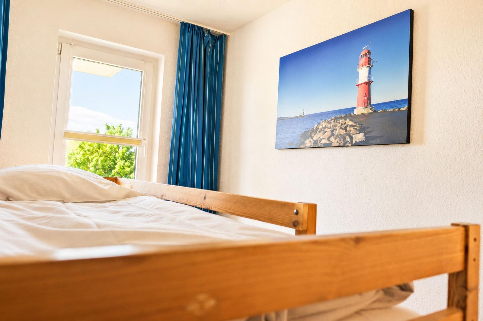 Ein Bett mit Holzrahmen, ein Fenster mit blauen Vorhängen und ein Bild eines Leuchtturms an der Wand.