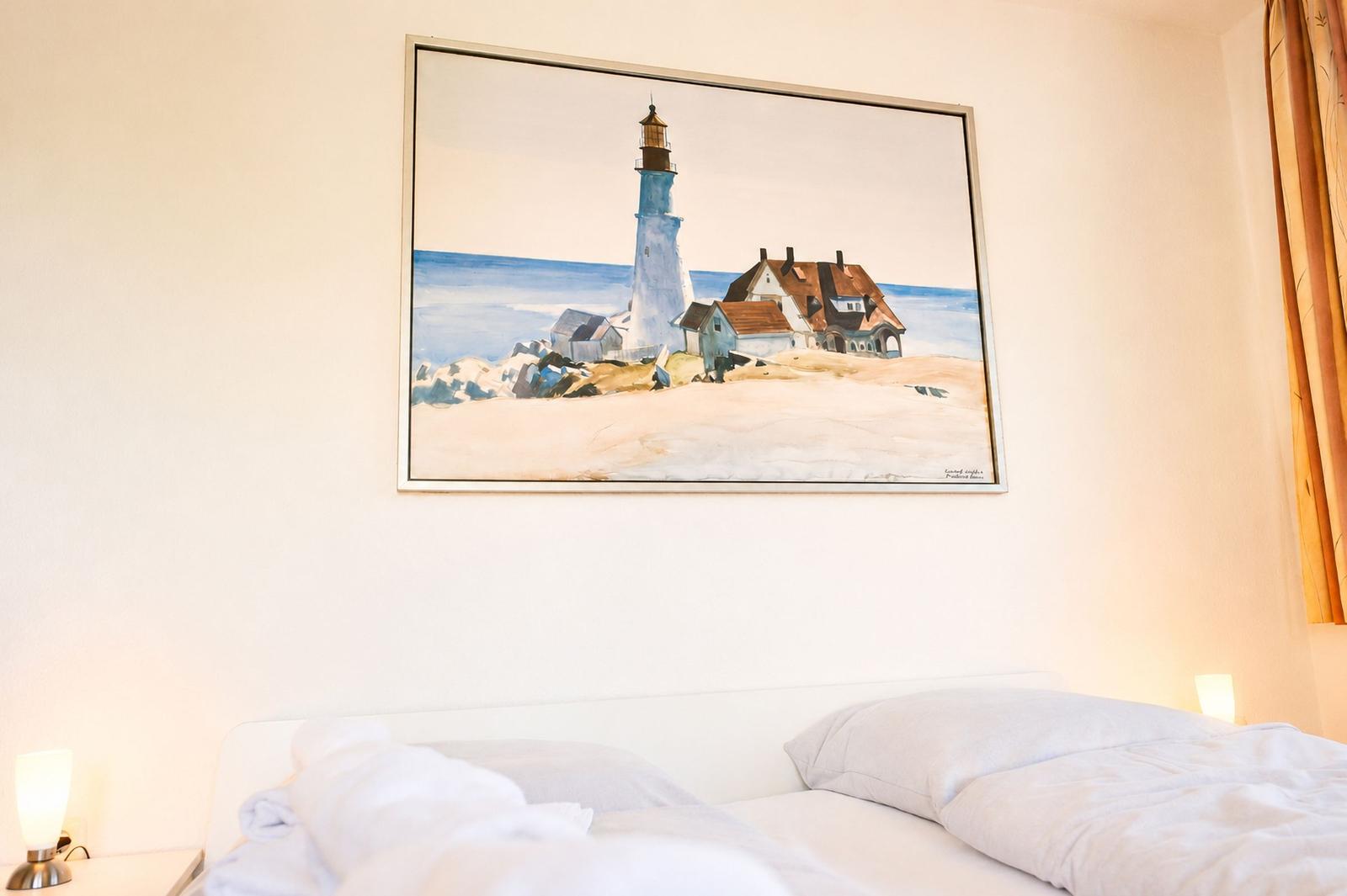Hinter dem Bett hängt ein Bild mit Leuchtturm und Häusern an der Küste.